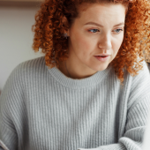 A woman with curly red hair, wearing a gray sweater, concentrates while writing with a pen at a desk
