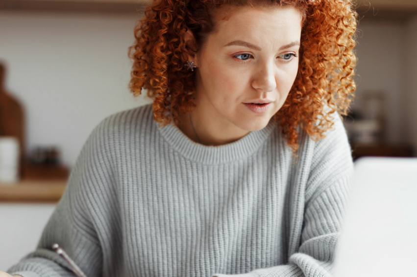 A woman with curly red hair, wearing a gray sweater, concentrates while writing with a pen at a desk