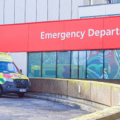Yellow and green ambulance parked outside an NHS hospital. The building has a large red sign reading 