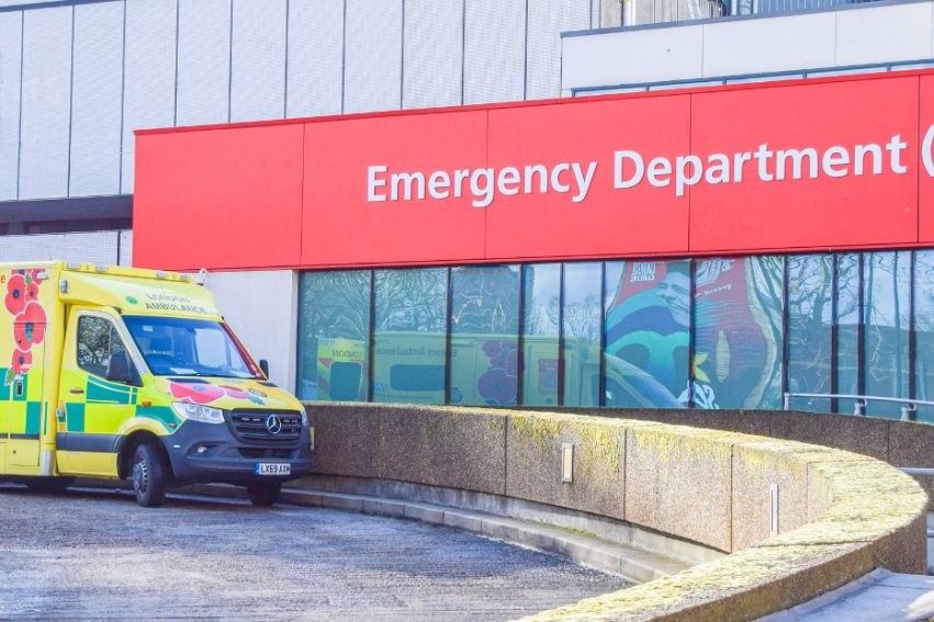 Yellow and green ambulance parked outside an NHS hospital. The building has a large red sign reading 