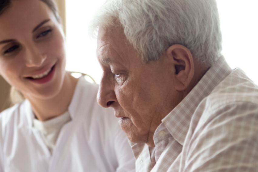 A smiling caregiver looks supportively at an elderly man with gray hair, who appears thoughtful