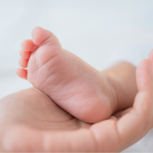 A gentle hand cradles a tiny baby foot, both resting on a soft white surface