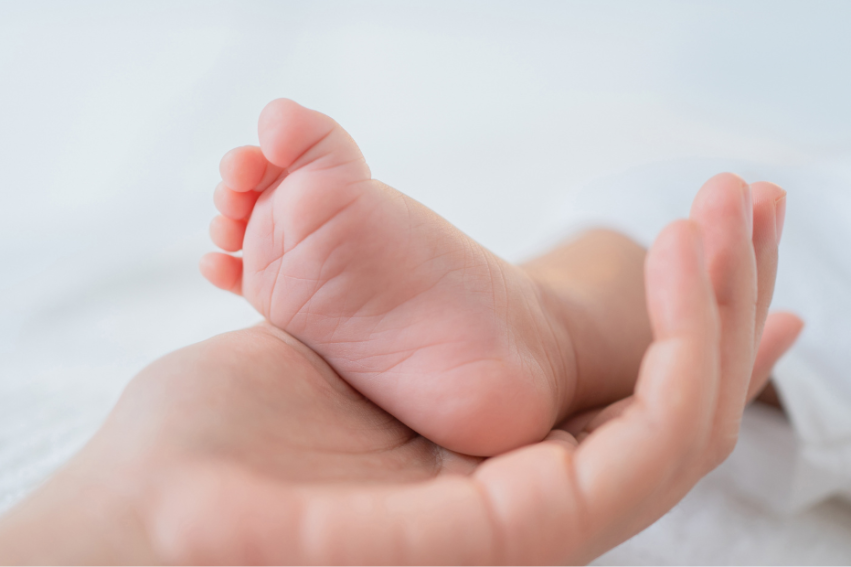 A gentle hand cradles a tiny baby foot, both resting on a soft white surface
