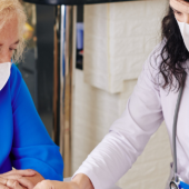 A healthcare professional in a white coat and face mask consults with an elderly woman in a blue shirt