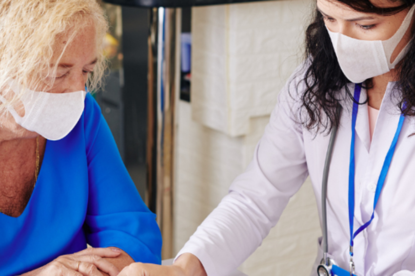 A healthcare professional in a white coat and face mask consults with an elderly woman in a blue shirt