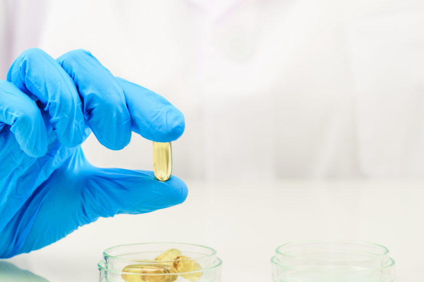 A person wearing blue gloves holds a clear capsule over an open petri dish containing similar capsules, set against a white laboratory backdrop.