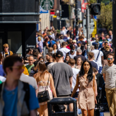 A bustling city street filled with pedestrians on a sunny day, with people of various ages and styles walking in both directions