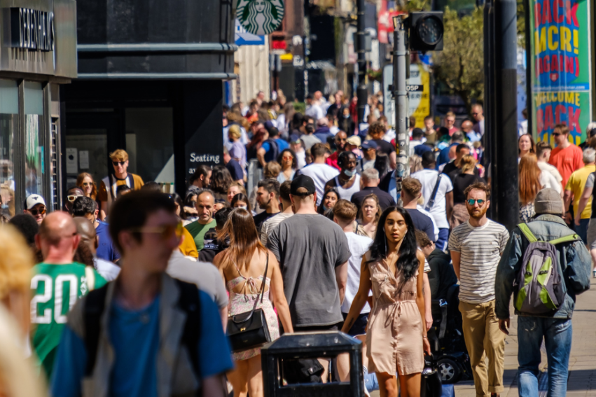 A bustling city street filled with pedestrians on a sunny day, with people of various ages and styles walking in both directions