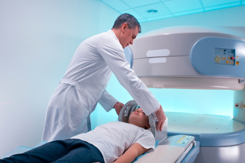A man doctor in a white coat adjusts a child patient's head for an MRI scan in a brightly lit room