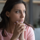 A woman in a pink top sits thoughtfully, hands clasped under her chin.