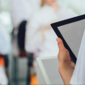 A person in a lab coat holds an electronic tablet or ipad, focusing on it, while blurred figures in similar attire sit in the background