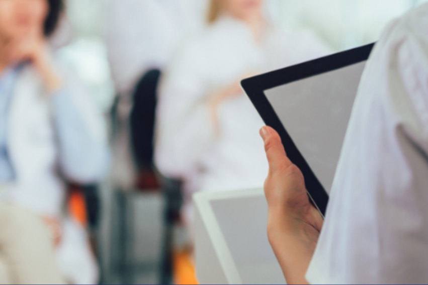 A person in a lab coat holds an electronic tablet or ipad, focusing on it, while blurred figures in similar attire sit in the background