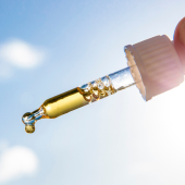 A hand holds a dropper filled with golden liquid against a bright sunny sky.