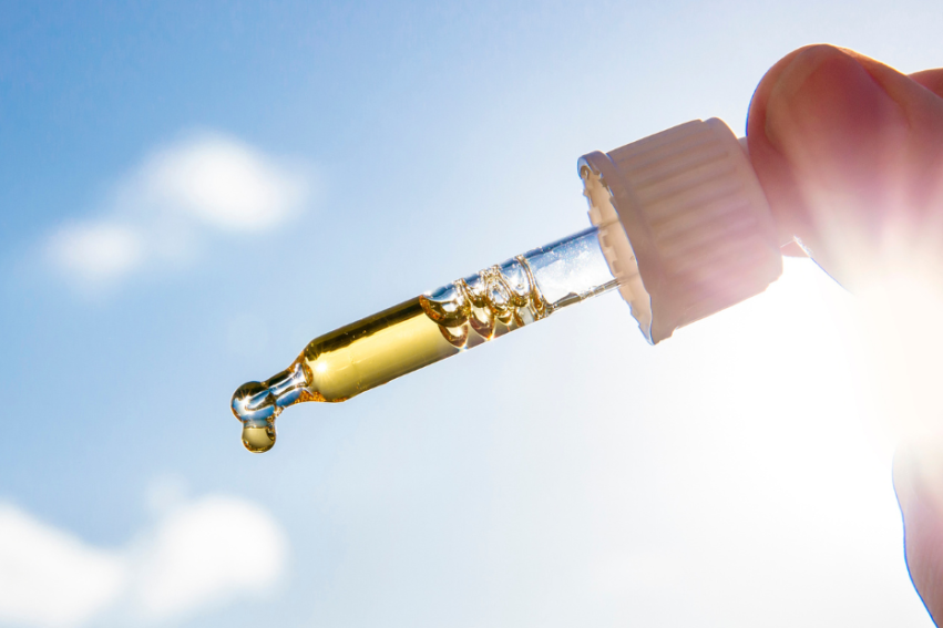 A hand holds a dropper filled with golden liquid against a bright sunny sky.