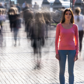 A woman in a pink sweater and jeans stands still on a busy street, surrounded by blurred pedestrians