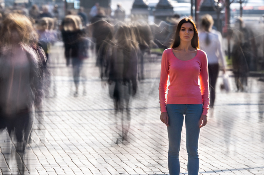 A woman in a pink sweater and jeans stands still on a busy street, surrounded by blurred pedestrians