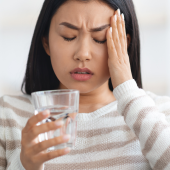 A woman in a striped sweater sits on a couch, holding her head with one hand and a glass of water in the other, appearing to have a headache.