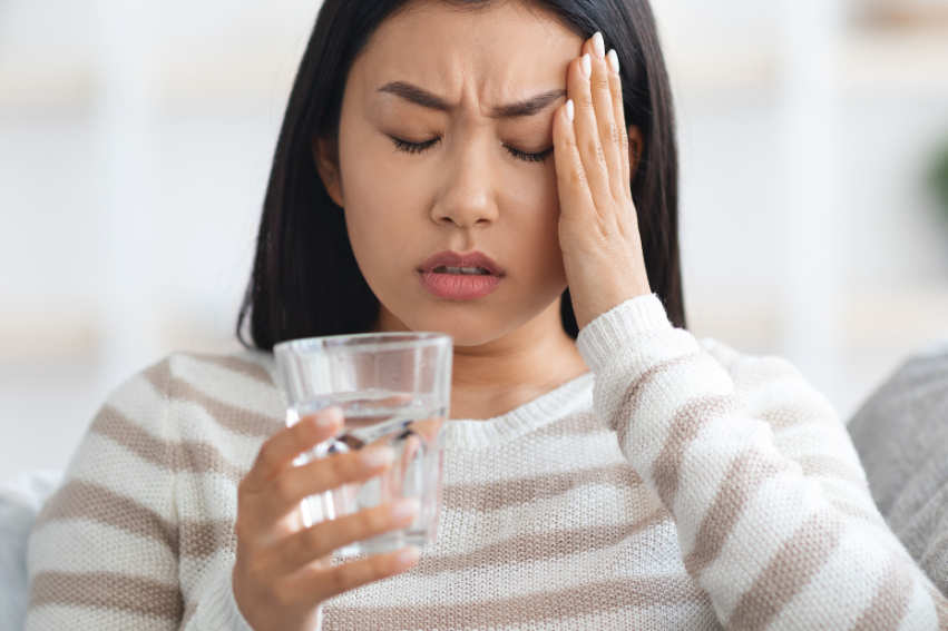 A woman in a striped sweater sits on a couch, holding her head with one hand and a glass of water in the other, appearing to have a headache.