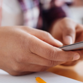 A person holds a smartphone in their hands above a notepad and pen. They wear a red smartwatch,