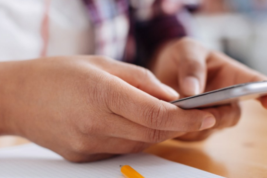 A person holds a smartphone in their hands above a notepad and pen. They wear a red smartwatch,
