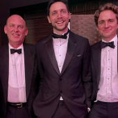 A group of six people dressed in formal attire, smiling. Three men in tuxedos with bow ties in the center, flanked by two women in floral dresses on each side.