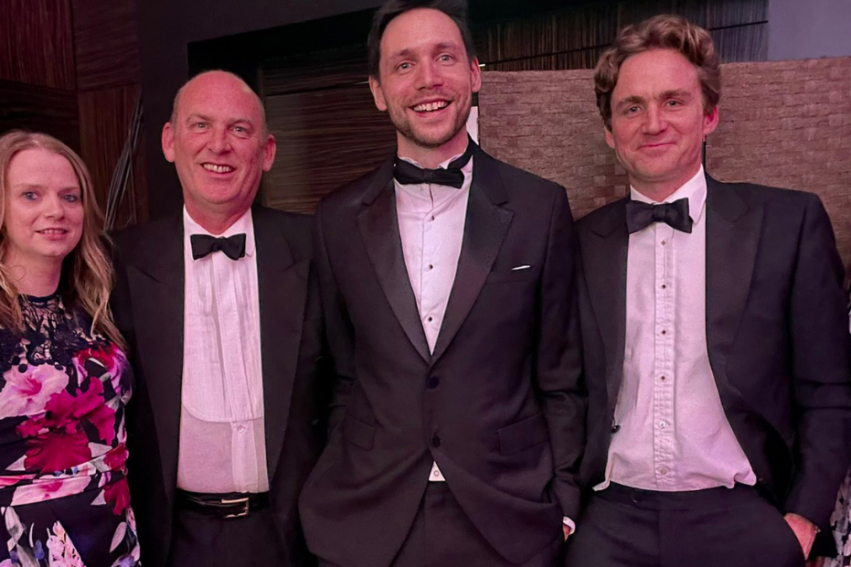 A group of six people dressed in formal attire, smiling. Three men in tuxedos with bow ties in the center, flanked by two women in floral dresses on each side.