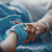 A healthcare worker in blue gloves adjusts an oximeter on a patient's finger in a hospital bed