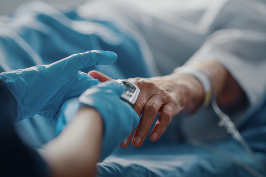 A healthcare worker in blue gloves adjusts an oximeter on a patient's finger in a hospital bed
