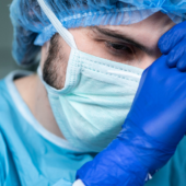 A healthcare worker in scrubs, mask, and gloves leans forward with clasped hands