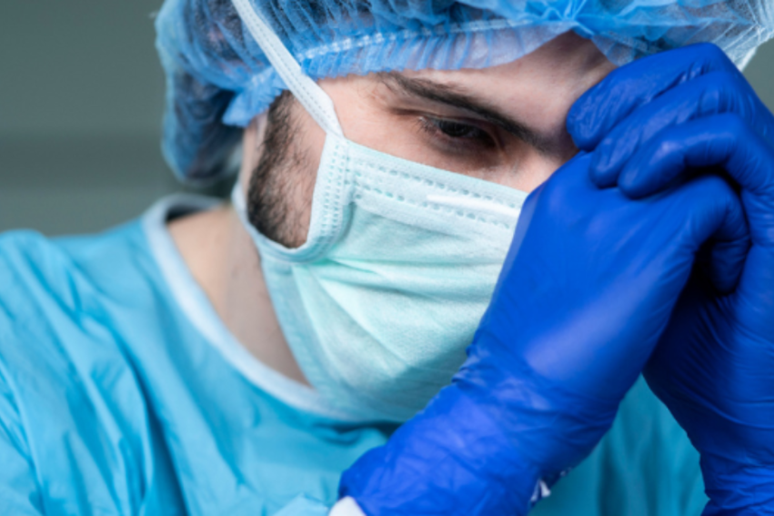 A healthcare worker in scrubs, mask, and gloves leans forward with clasped hands