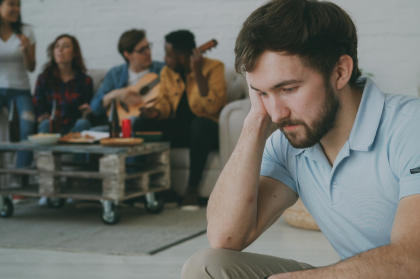 A man in the foreground appears sad and isolated at a social gathering