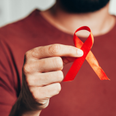 A person in a maroon shirt holds a red awareness ribbon between thumb and forefinger