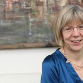 A smiling woman with short hair and glasses wears a blue blouse, seated on a white sofa.