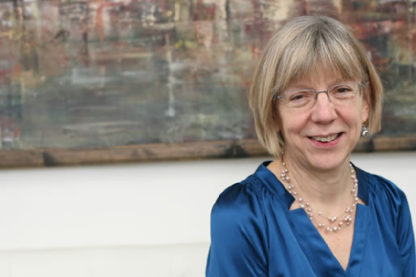 A smiling woman with short hair and glasses wears a blue blouse, seated on a white sofa.