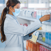 A female pharmacist in a white coat and face mask organizes medication boxes on pharmacy shelves