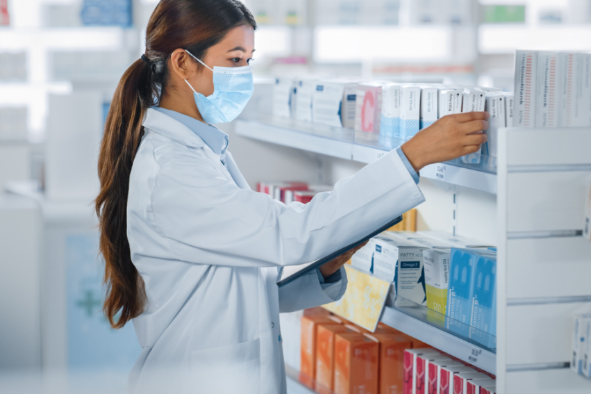 A female pharmacist in a white coat and face mask organizes medication boxes on pharmacy shelves
