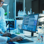 In a dimly lit lab, three scientists in white coats analyze data on a computer.