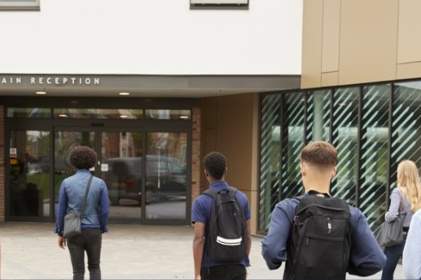 Students with backpacks walk towards a modern building's main reception entrance