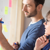 People collaborating at a whiteboard, writing notes on sticky notes. A man holds a marker, while a woman listens intently