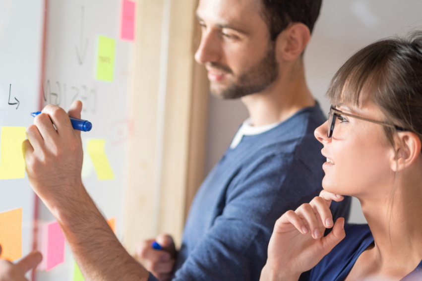 People collaborating at a whiteboard, writing notes on sticky notes. A man holds a marker, while a woman listens intently