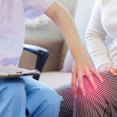 A healthcare professional in blue scrubs takes notes while a person in a white sweater touches their knee, highlighted with a red glow