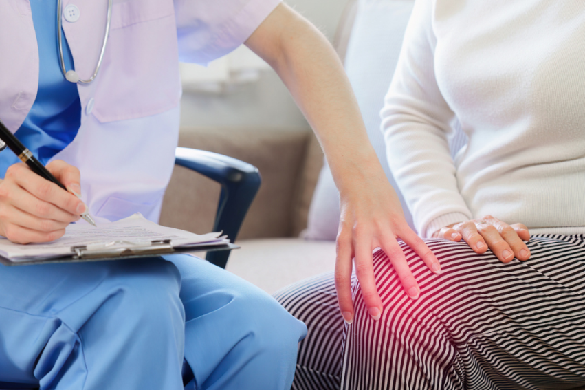 A healthcare professional in blue scrubs takes notes while a person in a white sweater touches their knee, highlighted with a red glow