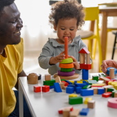 A child plays with colorful wooden blocks at a table, surrounded by smiling adults