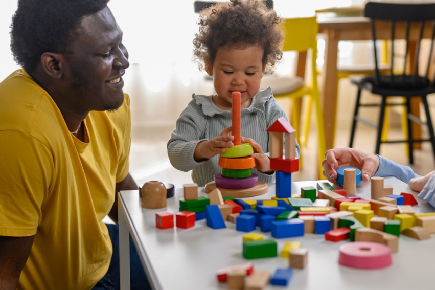 A child plays with colorful wooden blocks at a table, surrounded by smiling adults
