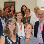 A group of smiling people, diverse in gender and attire, stand on a staircase, wearing badges