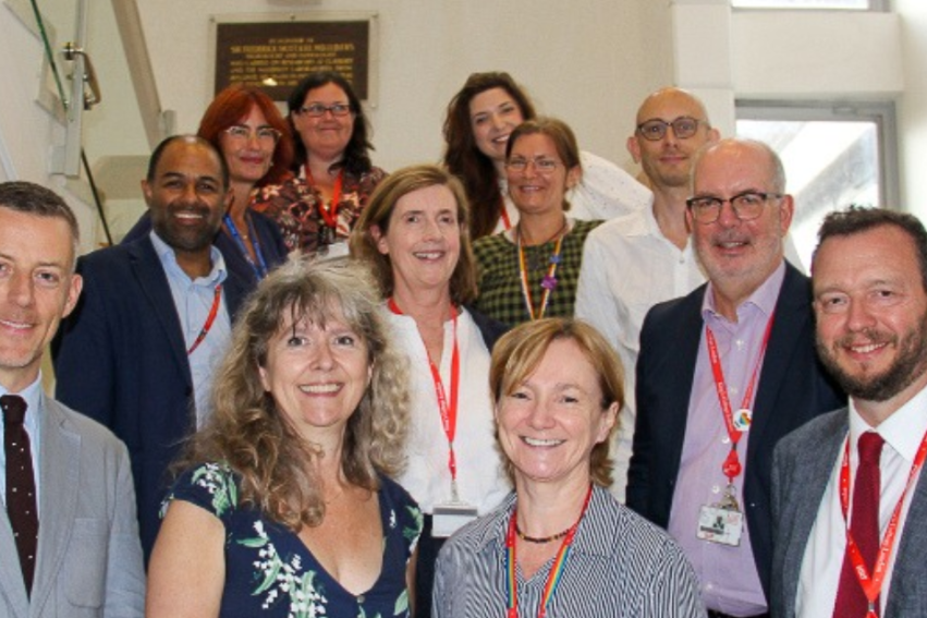 A group of smiling people, diverse in gender and attire, stand on a staircase, wearing badges
