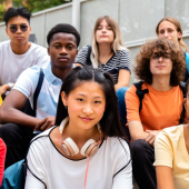 A diverse group of ten serious-looking teens sits on outdoor steps