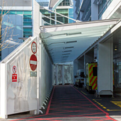 Modern hospital entrance with a covered driveway, red-lined road, and an ambulance. People walk nearby
