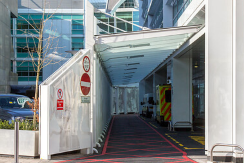 Modern hospital entrance with a covered driveway, red-lined road, and an ambulance. People walk nearby