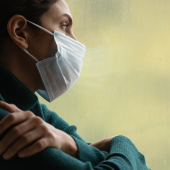 Woman in a mask looks out a window pensively. She wears a green sweater and rests her arms on each other